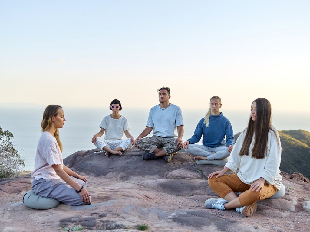 Five adults meditating outdoors on a mountain top during sunrise, promoting relaxation and mindfulness.